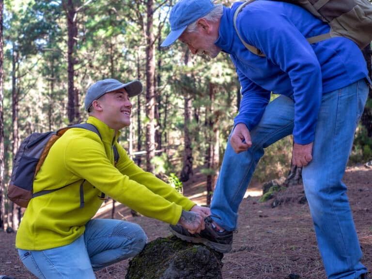 Young boy helps his grandfather to tie a shoe as they walk together in the mountain forest enjoying nature and healthy activity. The new generation helps the older one. Adventure has no age